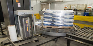 A pallet stacked with bags is being wrapped in plastic by an automated Wulftec stretch wrap machine in an industrial warehouse setting.