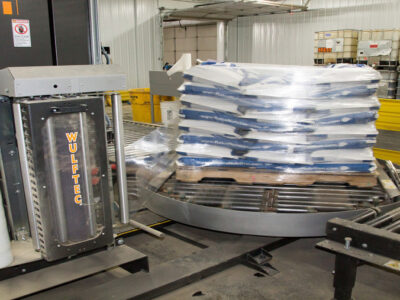 A pallet stacked with bags is being wrapped in plastic by an automated Wulftec stretch wrap machine in an industrial warehouse setting.