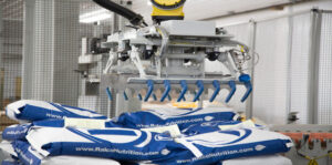 A robotic arm with a blue gripper is stacking large white and blue bags labeled "Ralco Nutrition" in a factory or warehouse setting.