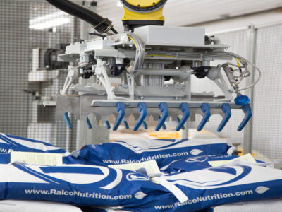 A robotic arm with a blue gripper is stacking large white and blue bags labeled "Ralco Nutrition" in a factory or warehouse setting.