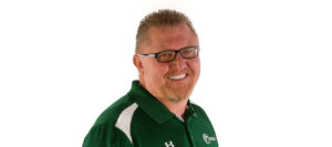 A smiling man with short spiked hair, glasses, and a goatee, wearing a green polo shirt with white accents and a logo, standing against a plain white background.