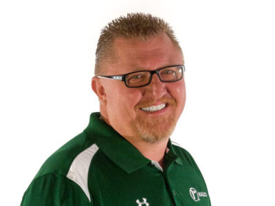 A smiling man with short spiked hair, glasses, and a goatee, wearing a green polo shirt with white accents and a logo, standing against a plain white background.