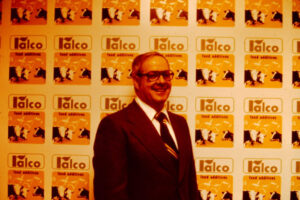 A man in a suit and tie stands smiling in front of a wall covered with repeated “Talco food additives” logos featuring cow graphics. The image has an orange tint.