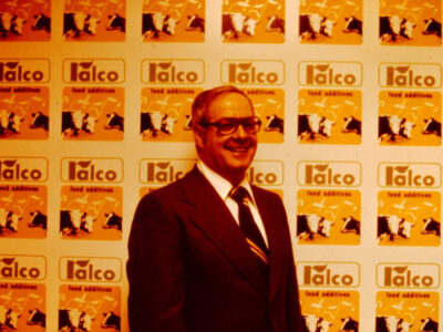 A man in a suit and tie stands smiling in front of a wall covered with repeated “Talco food additives” logos featuring cow graphics. The image has an orange tint.