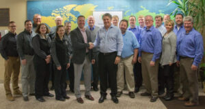 A group of about twenty people, mostly men and a few women, poses indoors in business casual attire. Two men stand in the center shaking hands, with a colorful world map displayed on the wall behind them.
