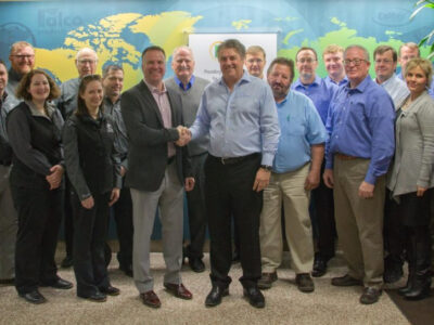 A group of about twenty people, mostly men and a few women, poses indoors in business casual attire. Two men stand in the center shaking hands, with a colorful world map displayed on the wall behind them.