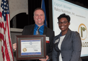 Two people stand smiling, one holding a framed certificate. An American flag is beside them. A screen in the background reads "Export Winner: Ralco Nutrition, Inc." with the company's logo displayed.