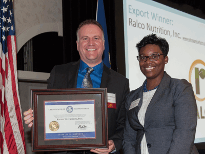 Two people stand smiling, one holding a framed certificate. An American flag is beside them. A screen in the background reads "Export Winner: Ralco Nutrition, Inc." with the company's logo displayed.