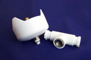 Two white plastic plumbing fittings with metal threads rest on a blue surface; one part is bowl-shaped and upright, the other is a T-shaped connector lying beside it.