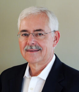 A man with short white hair, glasses, and a mustache is wearing a dark suit jacket over a white collared shirt, standing in front of a plain, light-colored background.