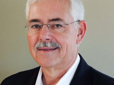 A man with short white hair, glasses, and a mustache is wearing a dark suit jacket over a white collared shirt, standing in front of a plain, light-colored background.