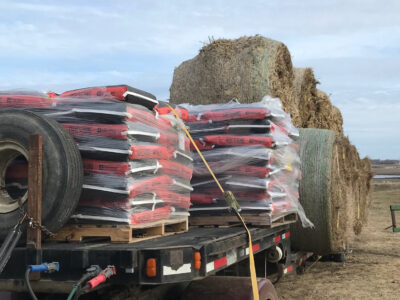A flatbed trailer loaded with stacked bags of feed wrapped in plastic and several large round hay bales sits on a farm under a cloudy sky. A spare tire is attached to the trailer.