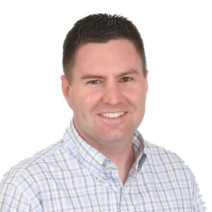 Smiling man with short dark hair wearing a light blue and white plaid button-up shirt, photographed against a plain white background.
