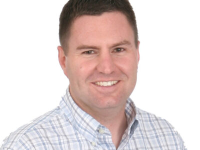 Smiling man with short dark hair wearing a light blue and white plaid button-up shirt, photographed against a plain white background.