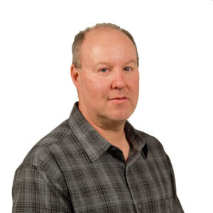A middle-aged man with short light hair and fair skin wearing a gray plaid button-up shirt, looking at the camera with a neutral expression against a white background.