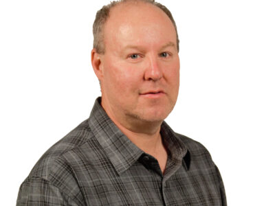 A middle-aged man with short light hair and fair skin wearing a gray plaid button-up shirt, looking at the camera with a neutral expression against a white background.
