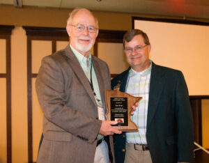 Two older men stand indoors smiling at the camera. One is holding a wooden state-shaped award plaque. Both are dressed in jackets and collared shirts, and a projection screen is visible in the background.
