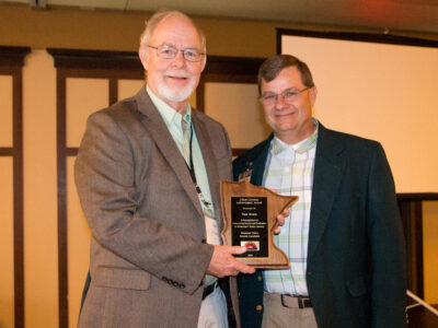 Two older men stand indoors smiling at the camera. One is holding a wooden state-shaped award plaque. Both are dressed in jackets and collared shirts, and a projection screen is visible in the background.