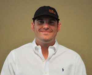 A man in a white button-down shirt and black cap stands against a plain tan background, smiling at the camera. The cap has orange text and the shirt has a small blue logo on the chest.
