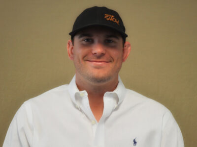 A man in a white button-down shirt and black cap stands against a plain tan background, smiling at the camera. The cap has orange text and the shirt has a small blue logo on the chest.