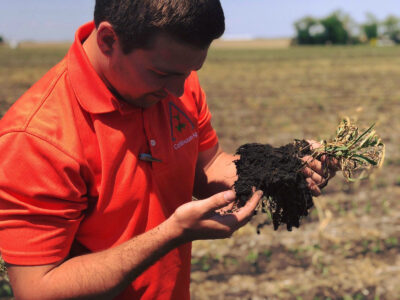 A man in an orange shirt examines soil and roots from a plant in a field, closely inspecting the quality and texture of the earth in his hands.