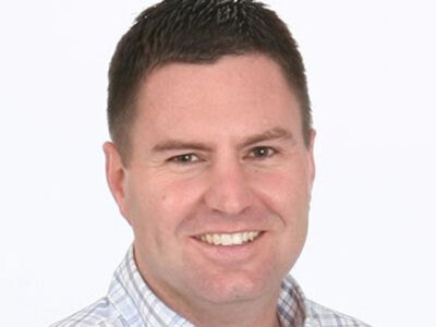 A man with short dark hair, wearing a light-colored, checkered shirt, smiles at the camera against a plain white background.