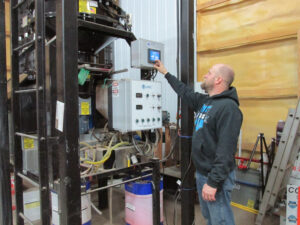 A man in a hoodie operates a control panel mounted on industrial equipment inside a workshop with yellow insulation on the wall. Various wires, pipes, and barrels are visible around the machinery.
