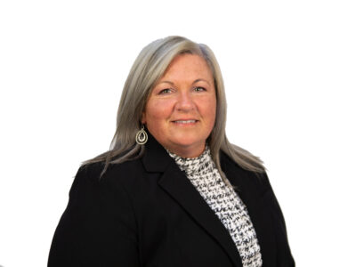 A woman with long gray hair wearing a black blazer and patterned top, smiling at the camera against a plain white background.