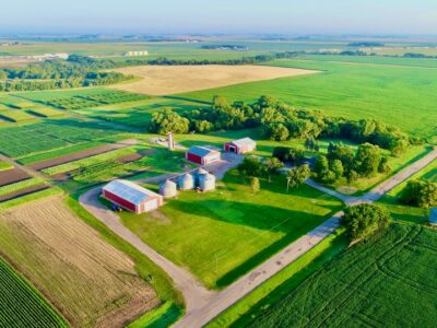 Aerial view of a large, green rural farm with red barns, silos, various fields, trees, and surrounding farmland under a clear blue sky.