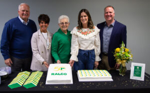 Five people stand behind a table with two decorated cakes, yellow flowers, plates, and napkins. The cake on the left has "50 RALCO" written on it, celebrating a 50th anniversary. All are smiling and dressed smartly.