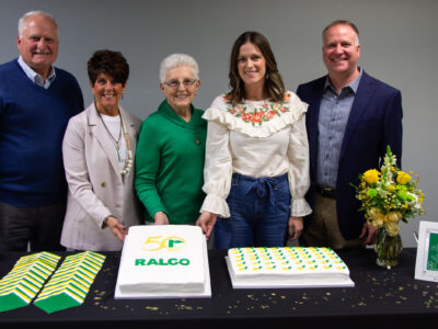 Five people stand behind a table with two decorated cakes, yellow flowers, plates, and napkins. The cake on the left has "50 RALCO" written on it, celebrating a 50th anniversary. All are smiling and dressed smartly.