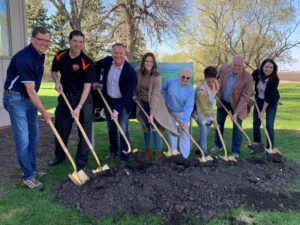Eight people stand in a row, each holding a shovel and smiling as they break ground on a pile of dirt outdoors, surrounded by green grass and trees on a sunny day.