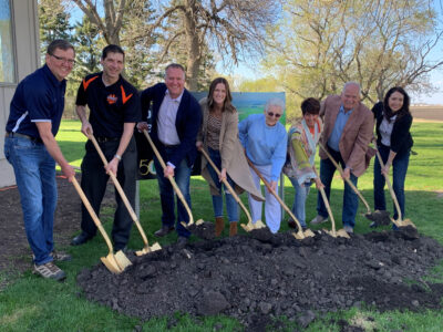 Eight people stand in a row, each holding a shovel and smiling as they break ground on a pile of dirt outdoors, surrounded by green grass and trees on a sunny day.