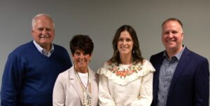Four adults stand side by side, smiling at the camera, in front of a plain gray wall. Two men are on the ends wearing jackets, and two women stand in the middle, one in a white blazer and one in a white floral top.
