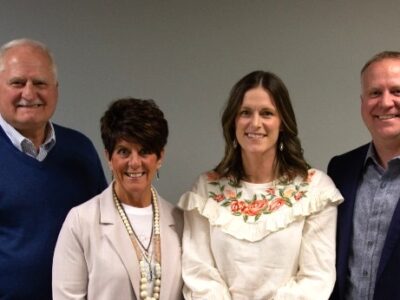 Four adults stand side by side, smiling at the camera, in front of a plain gray wall. Two men are on the ends wearing jackets, and two women stand in the middle, one in a white blazer and one in a white floral top.