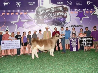 A group of people pose with a steer and awards at the Show Steer Junior Nationals. Signs, banners, a large check, and trophies are displayed. The background features logos, stars, and purple lighting.