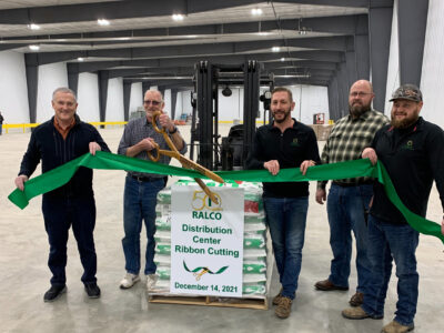 Five men smile while holding and cutting a large green ribbon in front of stacked pallets, marking the opening of a Ralco distribution center. A sign reads "Ribbon Cutting, December 14, 2021.