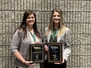 Two women stand side by side in front of a brick wall, smiling and holding award plaques. Both wear business attire and green lanyards around their necks.