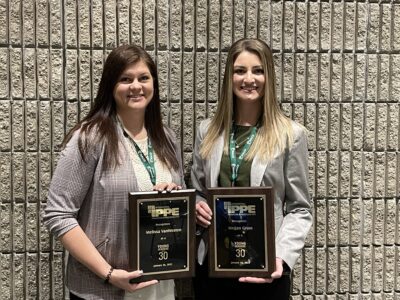 Two women stand side by side in front of a brick wall, smiling and holding award plaques. Both wear business attire and green lanyards around their necks.