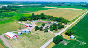 Aerial view of a rural farm with red barns, silver grain silos, scattered trees, and surrounding green and yellow fields under a clear blue sky. Dirt roads run alongside the property.
