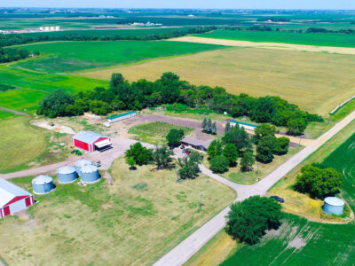 Aerial view of a rural farm with red barns, silver grain silos, scattered trees, and surrounding green and yellow fields under a clear blue sky. Dirt roads run alongside the property.