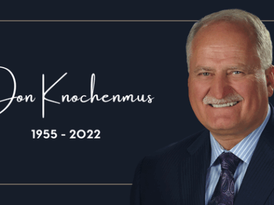 Memorial image with the text "Jon Knochenmus 1955 - 2022" next to a smiling older man in a suit and tie, set against a dark background with a simple border.