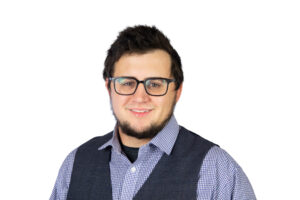 A young man with dark hair, a beard, and glasses is smiling. He is wearing a blue checkered shirt and a dark vest, standing against a plain white background.