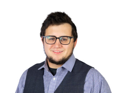 A young man with dark hair, a beard, and glasses is smiling. He is wearing a blue checkered shirt and a dark vest, standing against a plain white background.