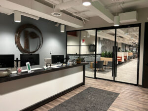 A modern office reception area with a white desk, computers, and a decorative round mirror on the wall. Beyond glass doors, there are open workspaces, plants, and lounge seating. The floor is wood with a rug.