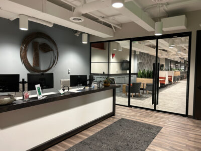 A modern office reception area with a white desk, computers, and a decorative round mirror on the wall. Beyond glass doors, there are open workspaces, plants, and lounge seating. The floor is wood with a rug.
