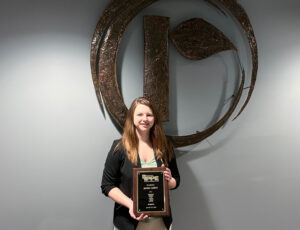A woman with long brown hair, wearing a black blazer, stands smiling and holding a plaque. She is in front of a large metallic logo on a gray wall.