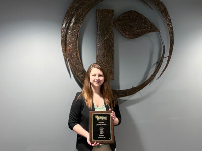 A woman with long brown hair, wearing a black blazer, stands smiling and holding a plaque. She is in front of a large metallic logo on a gray wall.
