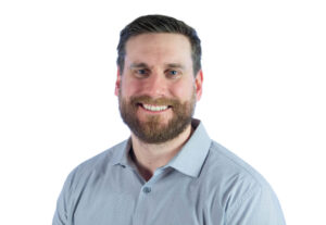A man with short brown hair and a beard smiles, wearing a light blue collared shirt, posed against a plain white background.