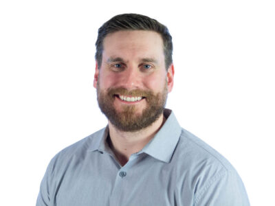 A man with short brown hair and a beard smiles, wearing a light blue collared shirt, posed against a plain white background.